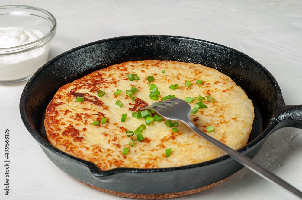 Swiss traditional dish is a potato pancake in a frying pan with a fork, decorated with green onions. there is a gravy boat with white cream sauce nearby. close-up.