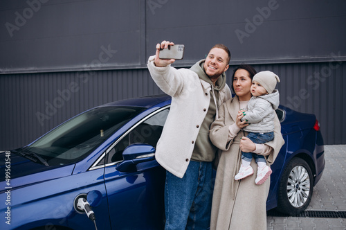 Beautiful young couple charges an electric car and takes a selfie on a smartphone