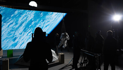 Behind the scenes of virtual production shot - Film crew working with Caucasian female astronaut stuntwoman in a spacesuit hanging on a wires against huge LED screen. Some elements furnished by NASA