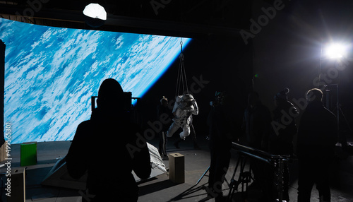 Fotografia Behind the scenes of virtual production shot - Film crew working with Caucasian female astronaut stuntwoman in a spacesuit hanging on a wires against huge LED screen