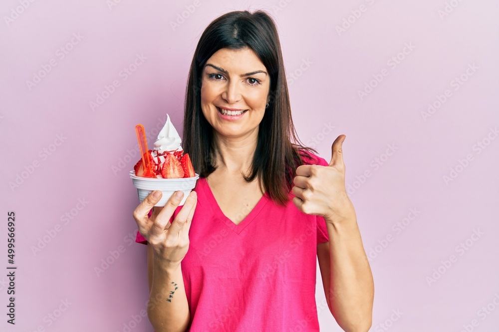 Young hispanic woman eating strawberry ice cream smiling happy and positive, thumb up doing excellent and approval sign