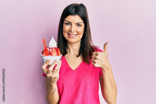 Young hispanic woman eating strawberry ice cream smiling happy and positive, thumb up doing excellent and approval sign