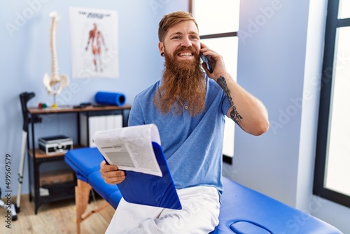Young redhead man wearing physiotherapist uniform talking on the smartphone at physiotherapy clinic