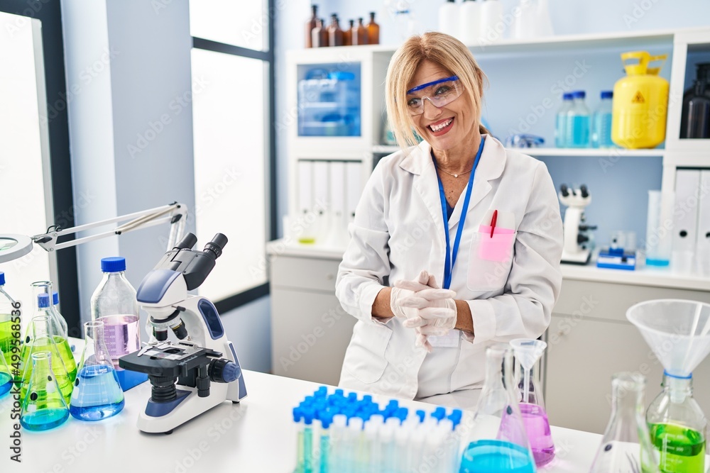 Middle age blonde woman working at scientist laboratory with hands together and crossed fingers smiling relaxed and cheerful. success and optimistic