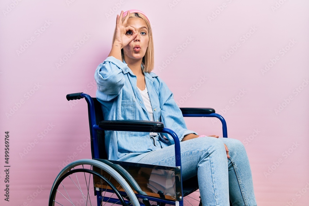 Beautiful blonde woman sitting on wheelchair doing ok gesture shocked with surprised face, eye looking through fingers. unbelieving expression.