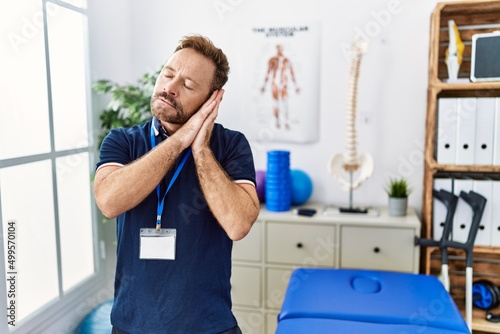 Middle age physiotherapist man working at pain recovery clinic sleeping tired dreaming and posing with hands together while smiling with closed eyes.