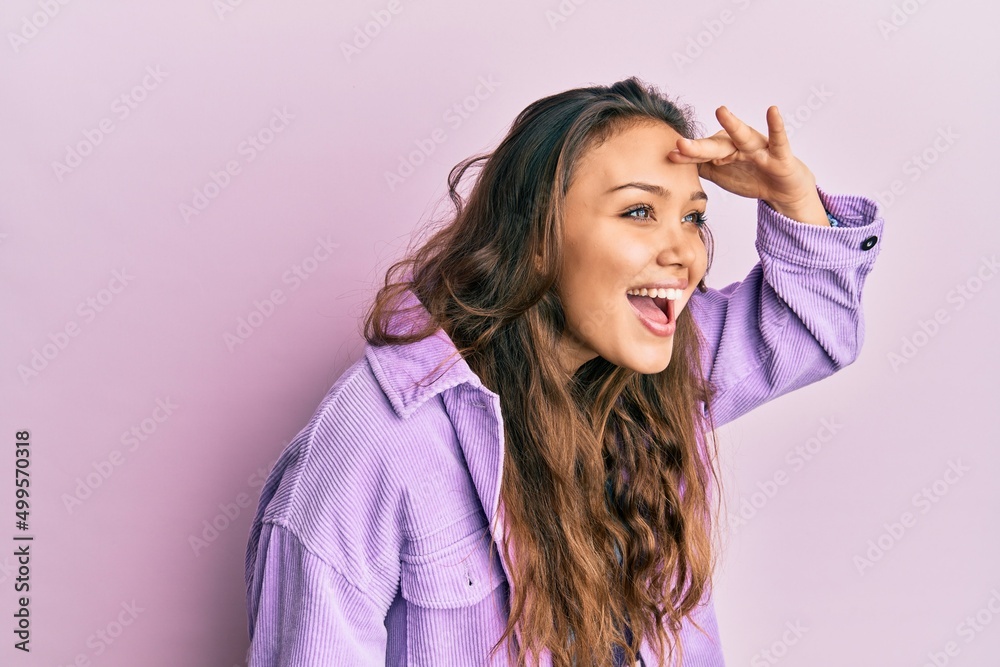 Young hispanic girl wearing casual clothes very happy and smiling looking far away with hand over head. searching concept. Young hispanic girl wearing casual clothes very happy and smiling looking far away with hand over head. searching concept.