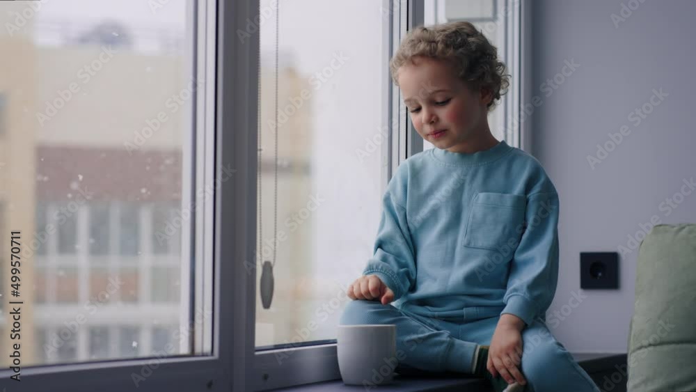 charming little boy is looking out window, sitting on still, watching falling snow in winter day