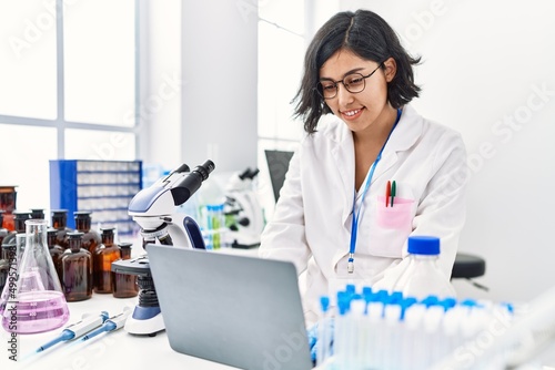 Young latin woman wearing scientist uniform using laptop working at laboratory