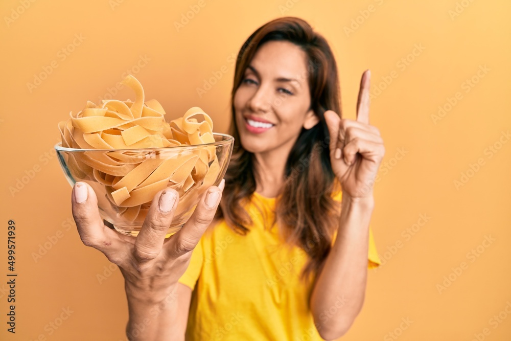Young latin woman holding bowl with uncooked pasta smiling happy pointing with hand and finger to the side Young latin woman holding bowl with uncooked pasta smiling happy pointing with hand and finger to the side