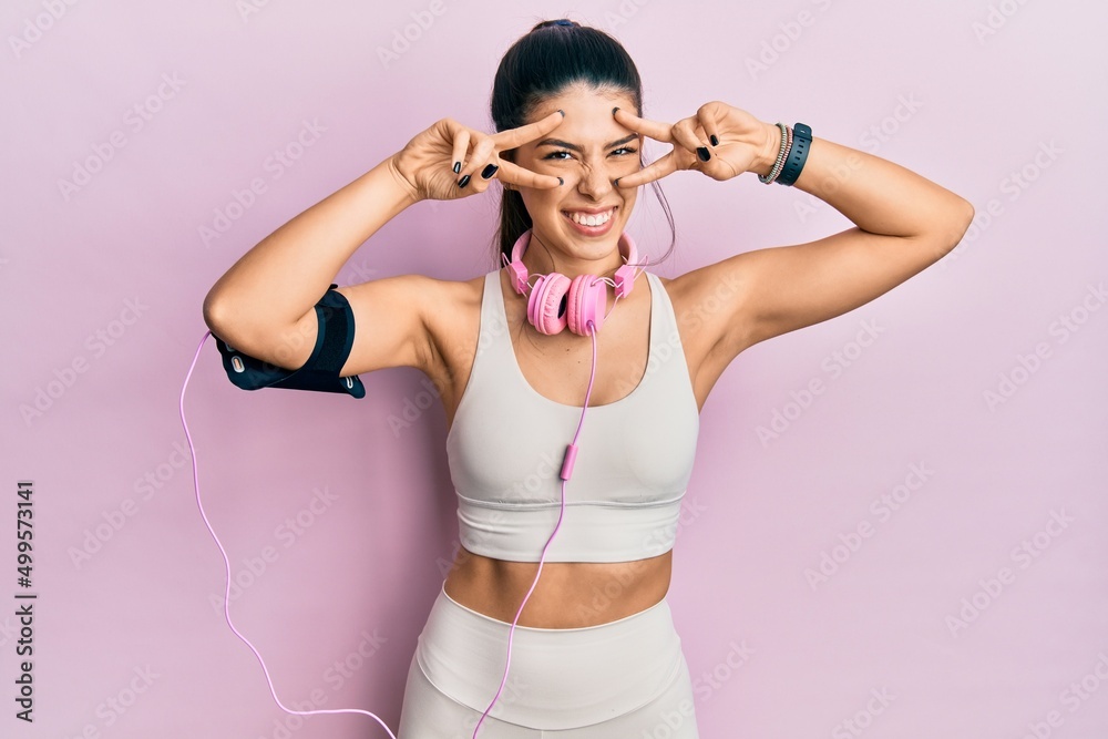 Young hispanic woman wearing gym clothes and using headphones doing peace symbol with fingers over face, smiling cheerful showing victory
