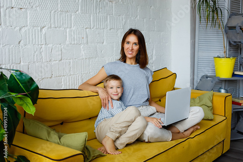 mother and son are sitting with a laptop and looking at the camera with a smile. a woman and a child on a yellow sofa.