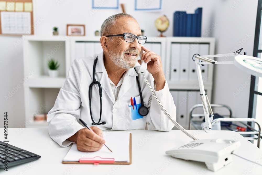 Senior grey-haired man wearing doctor uniform talking on the telephone at clinic Senior grey-haired man wearing doctor uniform talking on the telephone at clinic
