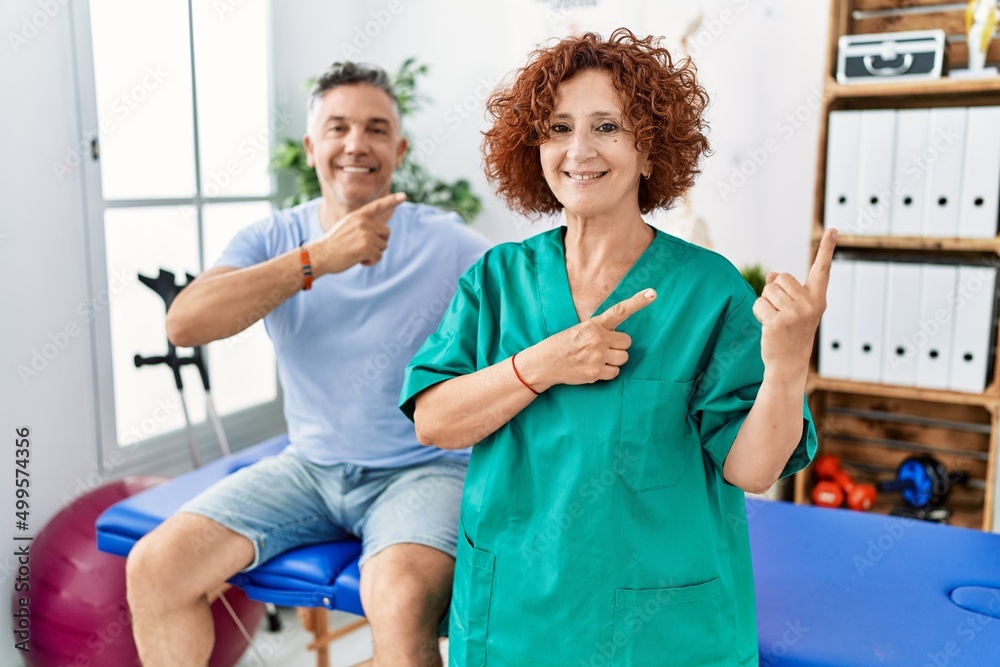 Obraz premium Physiotherapy woman working at pain recovery clinic with patient smiling and looking at the camera pointing with two hands and fingers to the side.