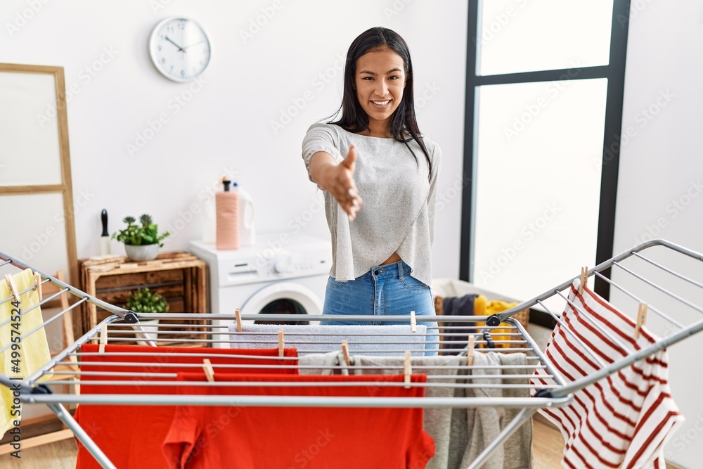 Obraz premium Young hispanic woman putting fresh laundry on clothesline smiling friendly offering handshake as greeting and welcoming. successful business.