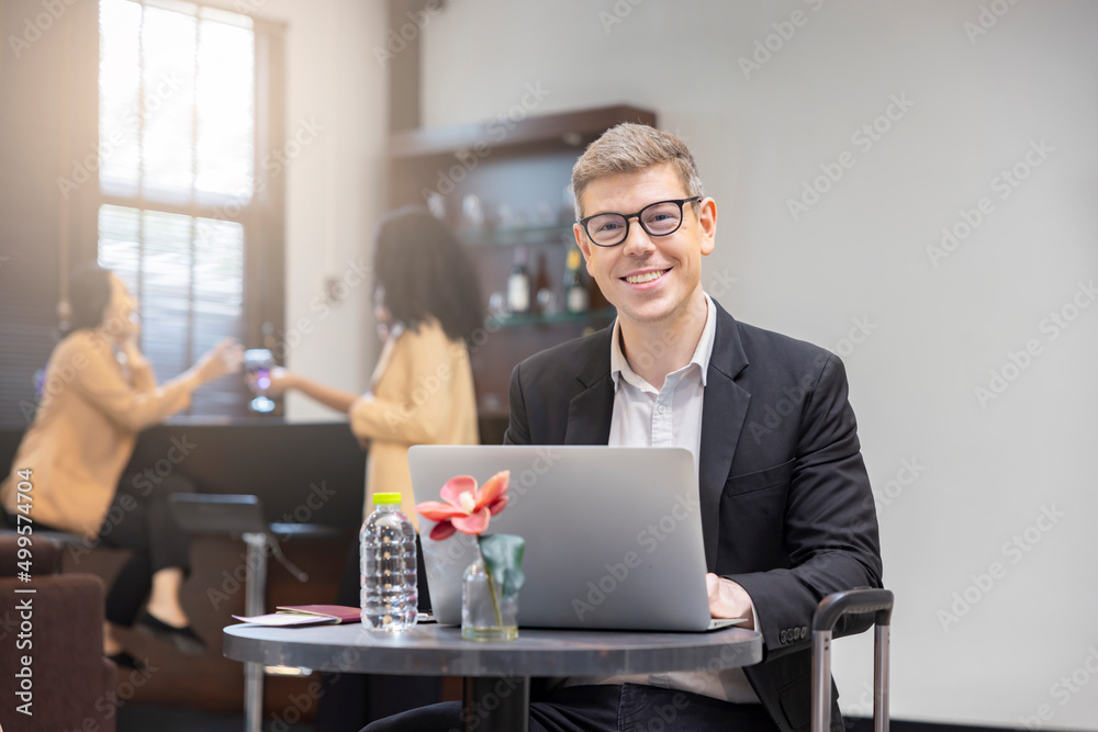 Smiling successful man entrepreneur in suite sitting at the airport lounge working on laptop. Passport, Boarding pass on table. Blurred background of passengers at counter serving drinks. Smiling successful man entrepreneur in suite sitting at the airport lounge working on laptop. Passport, Boarding pass on table. Blurred background of passengers at counter serving drinks.