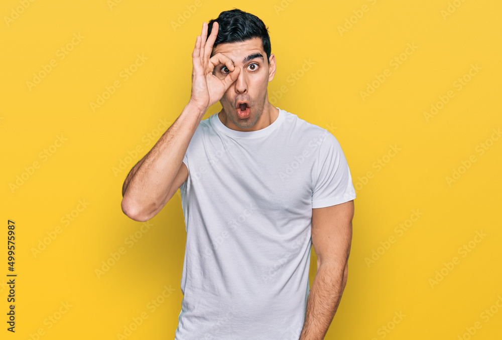 Handsome hispanic man wearing casual white t shirt doing ok gesture shocked with surprised face, eye looking through fingers. unbelieving expression. Handsome hispanic man wearing casual white t shirt doing ok gesture shocked with surprised face, eye looking through fingers. unbelieving expression.