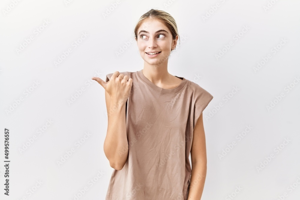 Young blonde woman standing over isolated background smiling with happy face looking and pointing to the side with thumb up. Young blonde woman standing over isolated background smiling with happy face looking and pointing to the side with thumb up.