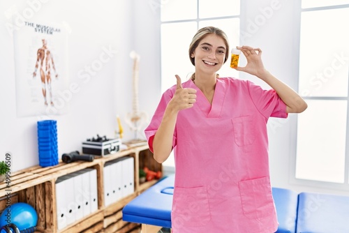 Young physiotherapist woman working at pain recovery clinic holding pills smiling happy and positive  thumb up doing excellent and approval sign