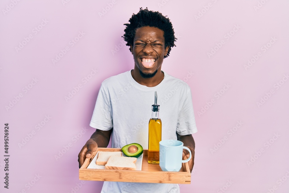 Young african american man holding tray with breakfast food sticking tongue out happy with funny expression. Young african american man holding tray with breakfast food sticking tongue out happy with funny expression.