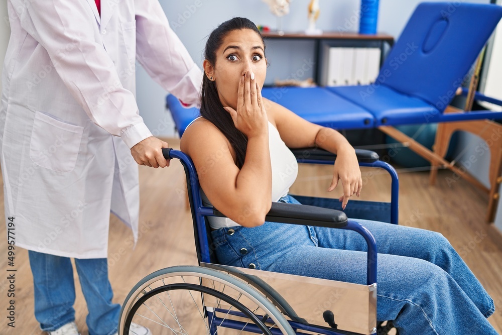 Obraz premium Young hispanic woman sitting on wheelchair at physiotherapy clinic covering mouth with hand, shocked and afraid for mistake. surprised expression