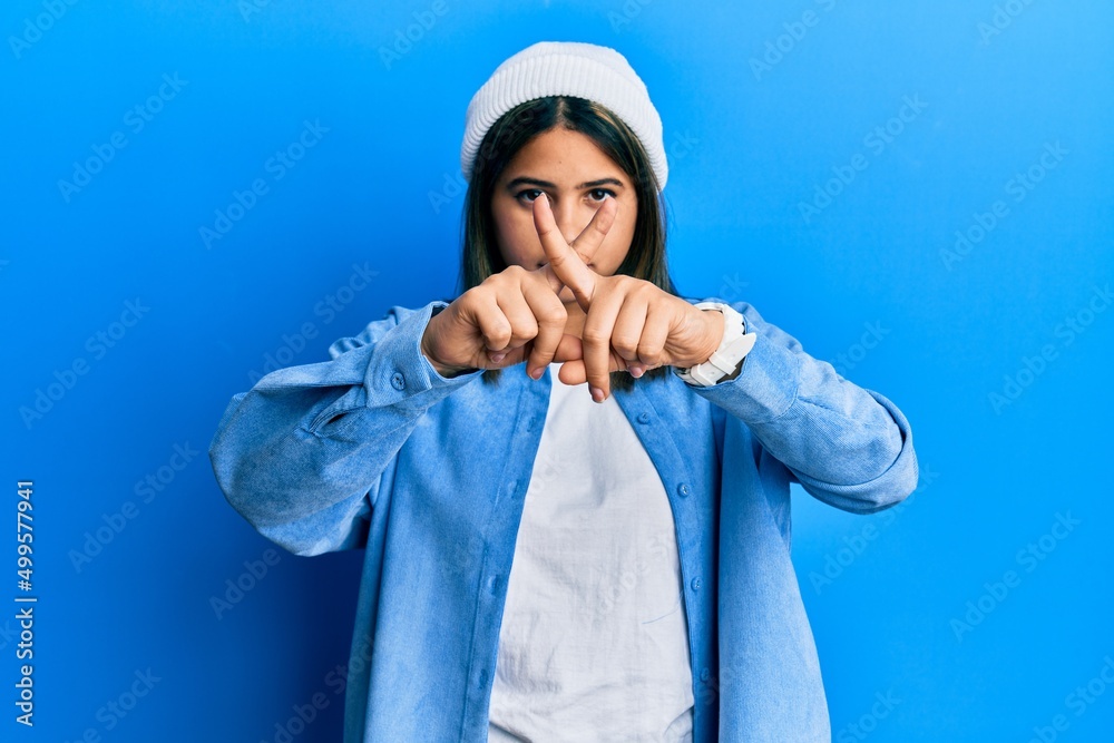 Young latin woman wearing cute wool cap rejection expression crossing fingers doing negative sign Young latin woman wearing cute wool cap rejection expression crossing fingers doing negative sign