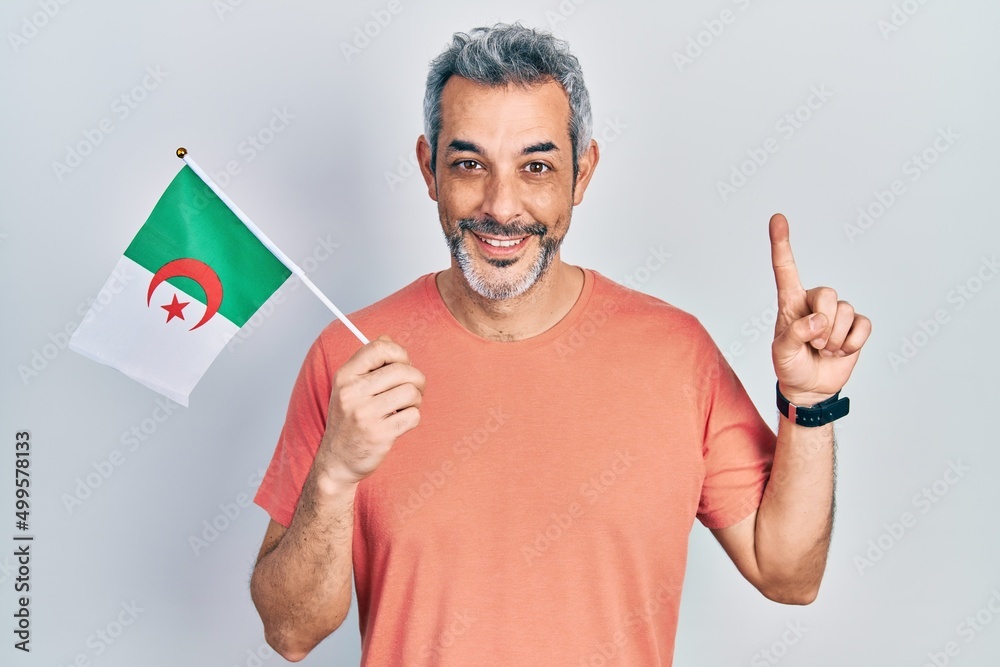 Handsome middle age man with grey hair holding algeria flag smiling with an idea or question pointing finger with happy face, number one Handsome middle age man with grey hair holding algeria flag smiling with an idea or question pointing finger with happy face, number one