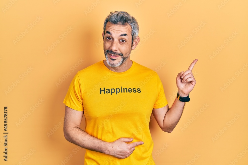 Handsome middle age man with grey hair wearing t shirt with happiness word message smiling happy pointing with hand and finger to the side Handsome middle age man with grey hair wearing t shirt with happiness word message smiling happy pointing with hand and finger to the side