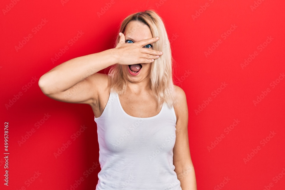 Beautiful caucasian blonde woman wearing casual white t shirt peeking in shock covering face and eyes with hand, looking through fingers with embarrassed expression. Beautiful caucasian blonde woman wearing casual white t shirt peeking in shock covering face and eyes with hand, looking through fingers with embarrassed expression.