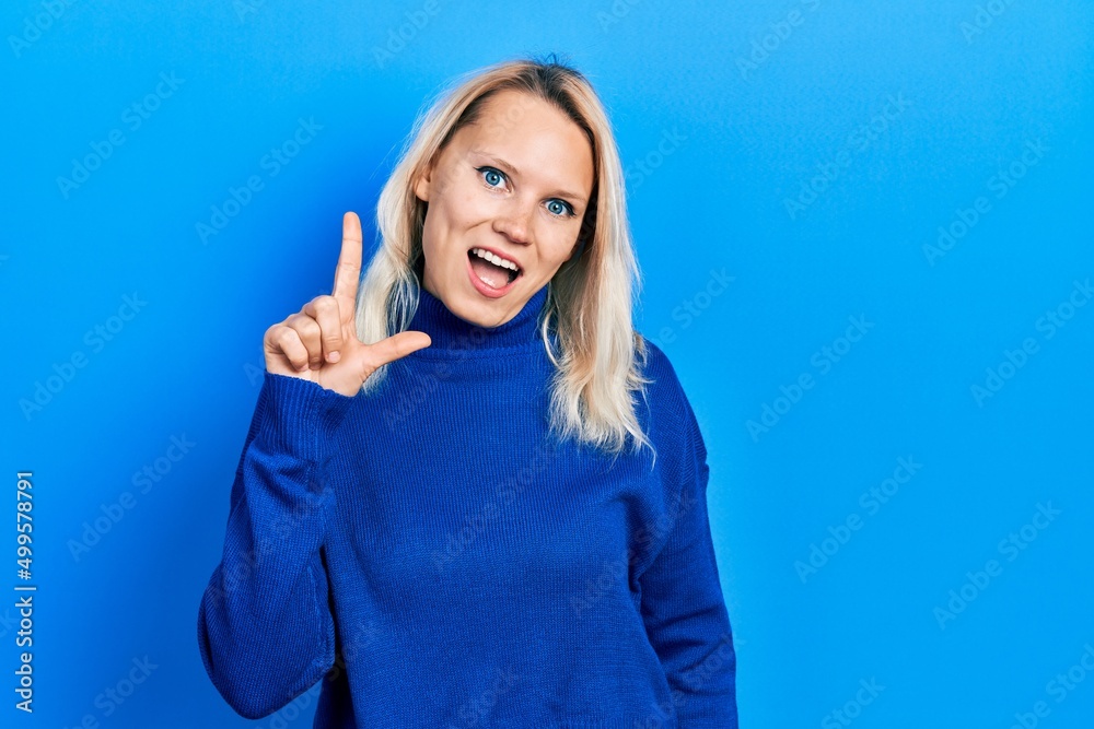 Beautiful caucasian blonde woman wearing turtleneck sweater smiling with an idea or question pointing finger up with happy face, number one