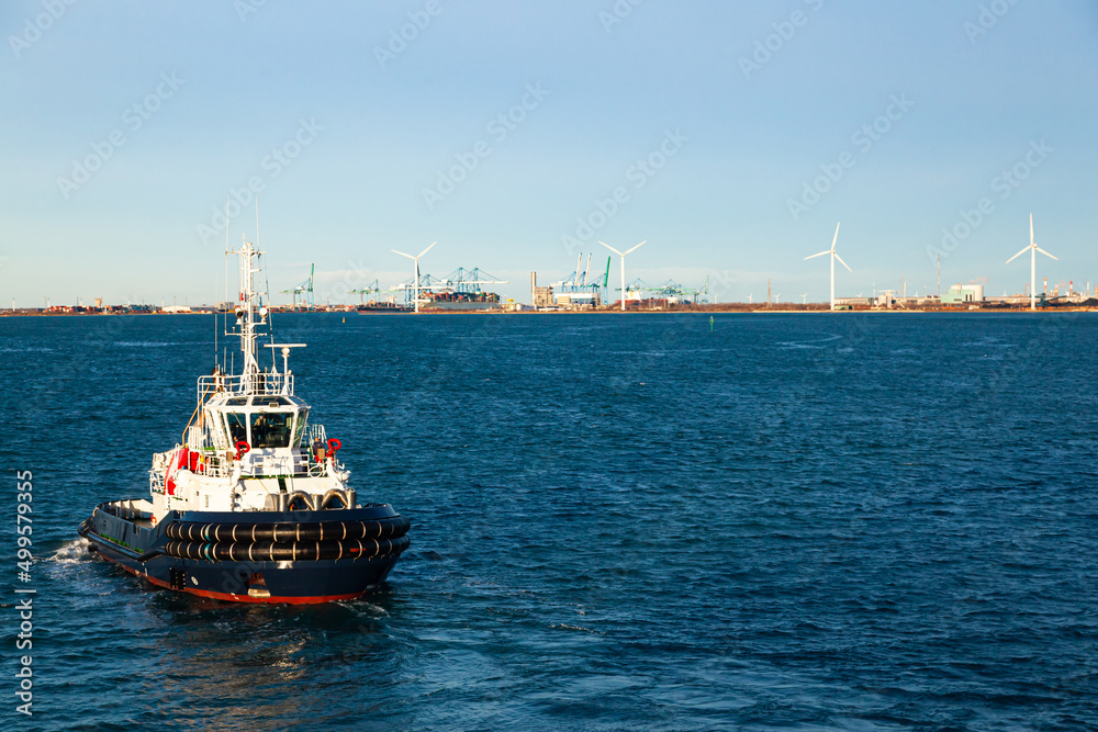 A tug boat sailing in the bay of the seaport of Fos-sur-Mer, France, with cranes and wind generators on the coast.
