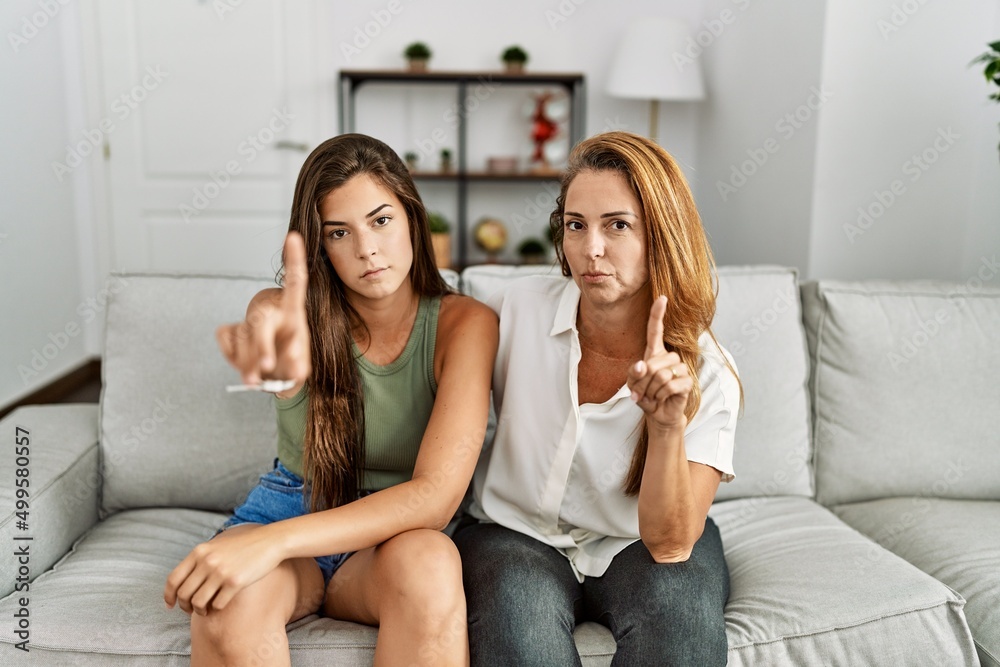 Mother and daughter together sitting on the sofa at home pointing with finger up and angry expression, showing no gesture Mother and daughter together sitting on the sofa at home pointing with finger up and angry expression, showing no gesture