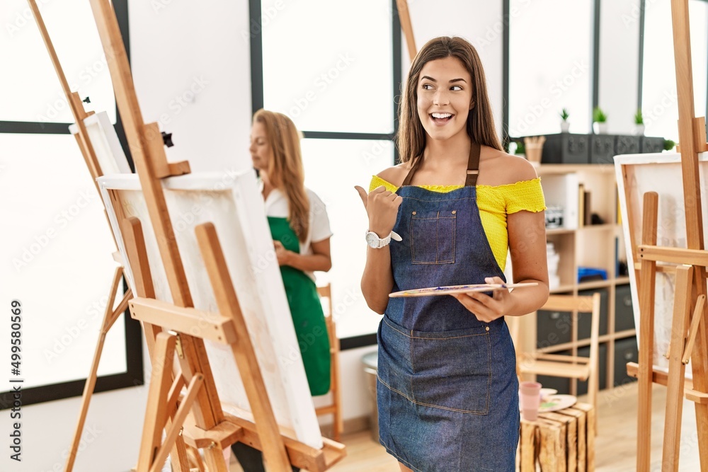 Obraz premium Young hispanic woman at art classroom pointing thumb up to the side smiling happy with open mouth