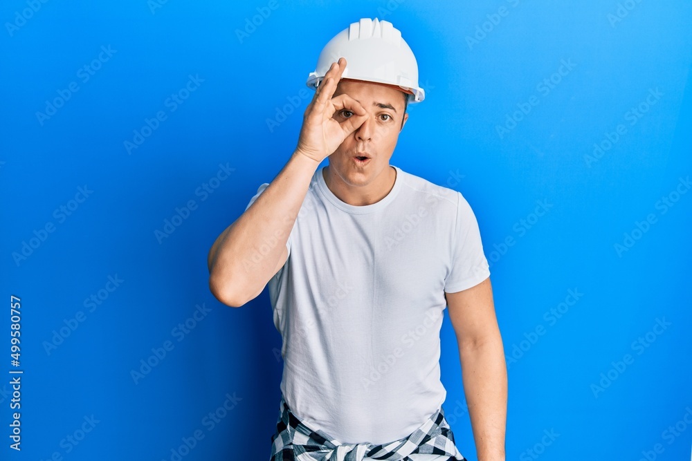 Handsome young man wearing builder uniform and hardhat doing ok gesture shocked with surprised face, eye looking through fingers. unbelieving expression.
