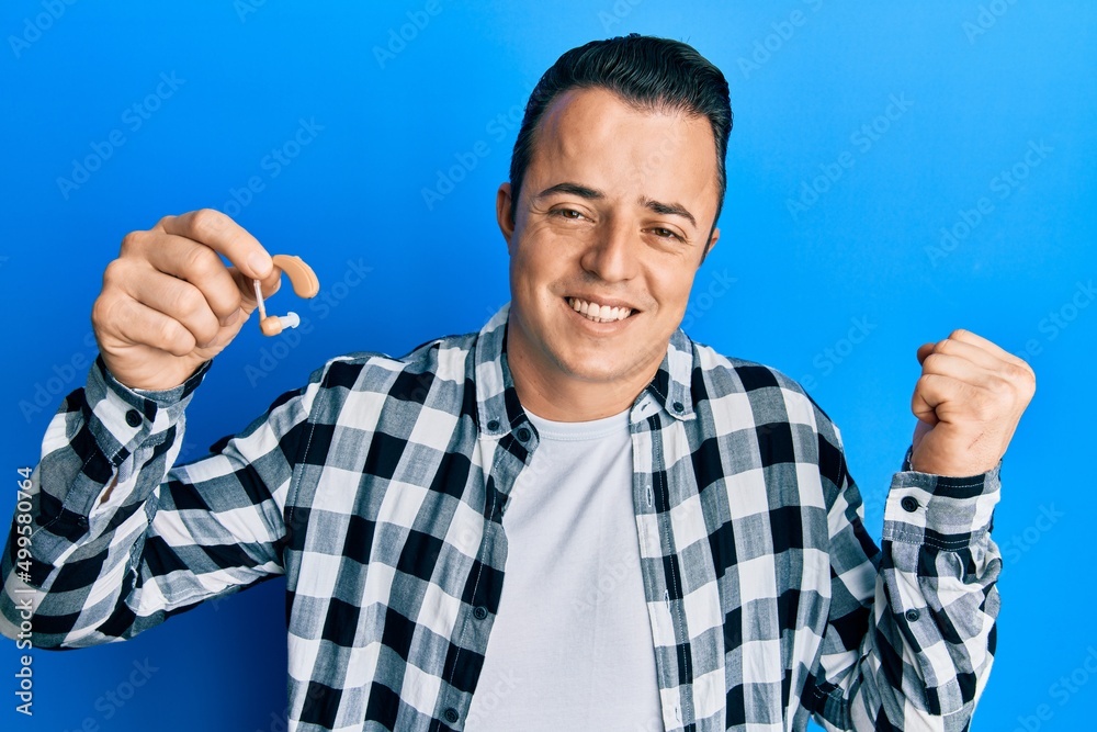Handsome young man holding medical hearing aid screaming proud, celebrating victory and success very excited with raised arm Handsome young man holding medical hearing aid screaming proud, celebrating victory and success very excited with raised arm