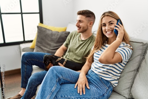 Young hispanic couple smiling happy talking on the smartphone sitting on the sofa with dog at home.