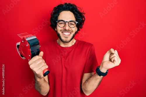Handsome hispanic man holding packing tape screaming proud, celebrating victory and success very excited with raised arm