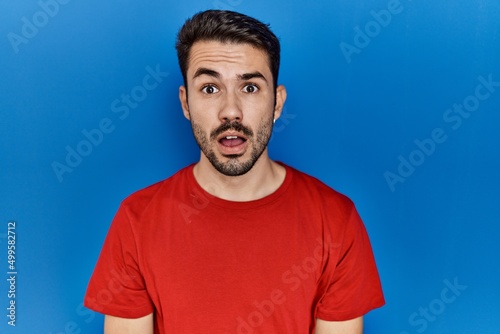 Young hispanic man with beard wearing red t shirt over blue background afraid and shocked with surprise expression, fear and excited face.