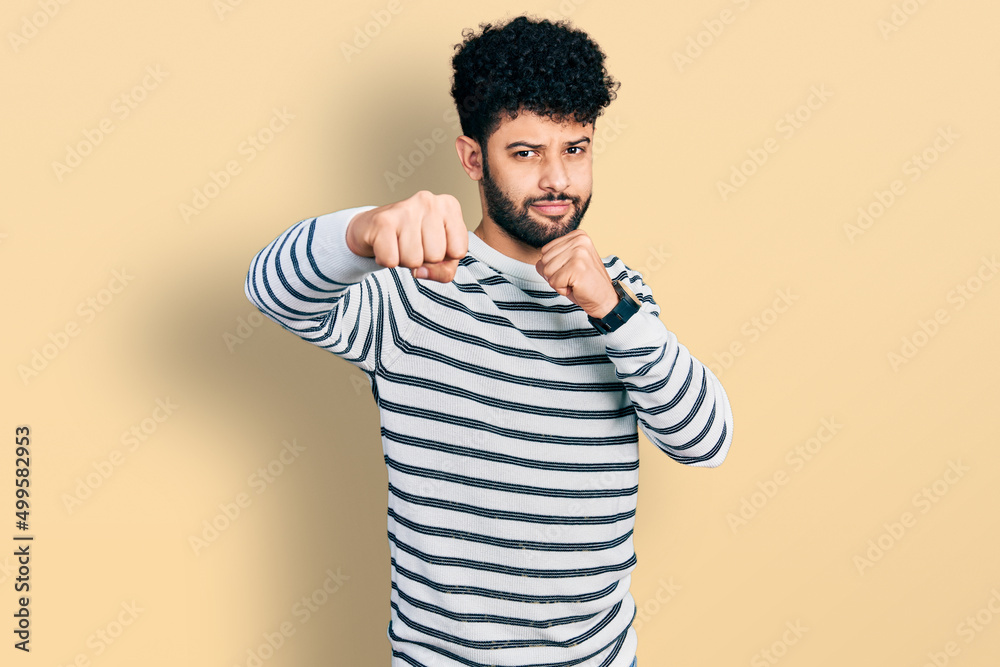 Young arab man with beard wearing casual striped sweater punching fist to fight, aggressive and angry attack, threat and violence Young arab man with beard wearing casual striped sweater punching fist to fight, aggressive and angry attack, threat and violence