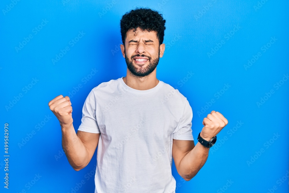 Young arab man with beard wearing casual white t shirt very happy and excited doing winner gesture with arms raised, smiling and screaming for success. celebration concept. Young arab man with beard wearing casual white t shirt very happy and excited doing winner gesture with arms raised, smiling and screaming for success. celebration concept.
