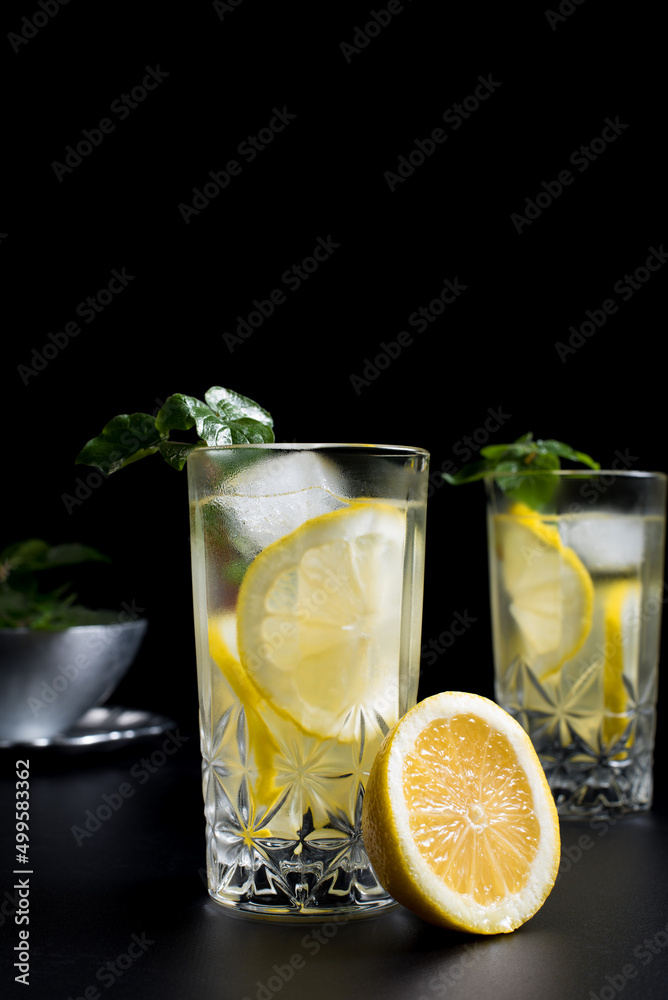 Fototapeta premium Dos vasos de refrescante zumo de limón, cóctel de frutas con rodajas de limón y hielo sobre fondo negro