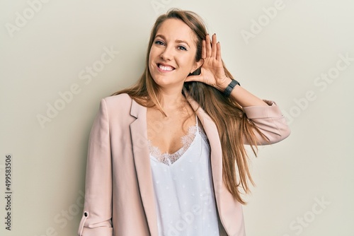 Young blonde woman wearing business jacket and glasses smiling with hand over ear listening an hearing to rumor or gossip. deafness concept.