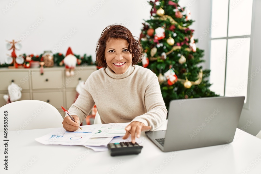 Middle age hispanic woman using laptop sitting by christmas tree at home.