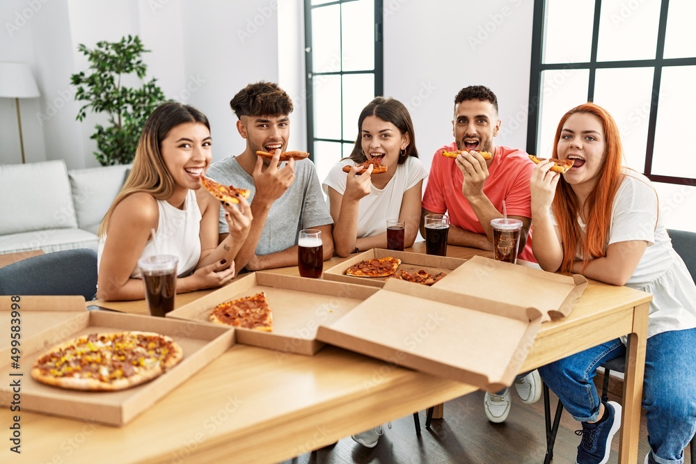 Group of young people smiling happy eating italian pizza sitting on the table at home Group of young people smiling happy eating italian pizza sitting on the table at home