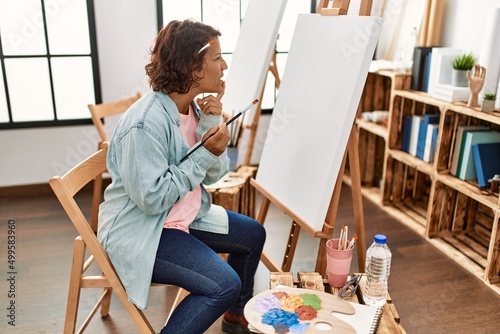 Middle age hispanic woman at art studio painting on canvas serious face thinking about question with hand on chin, thoughtful about confusing idea