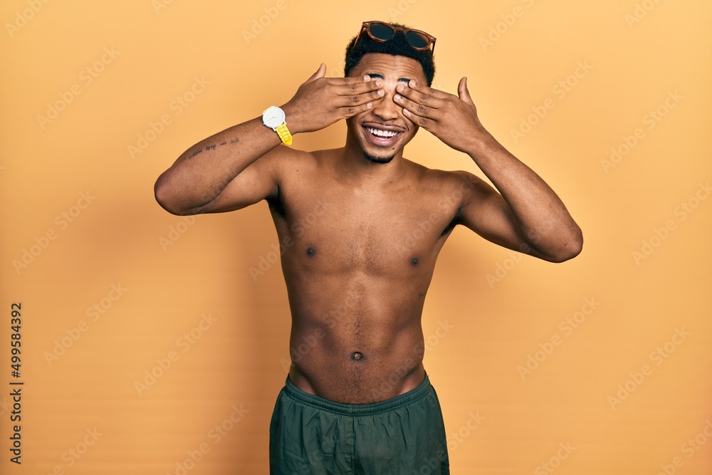Young african american man wearing swimwear and swimmer glasses covering eyes with hands smiling cheerful and funny. blind concept.