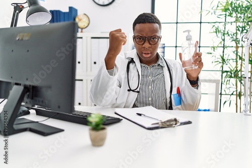 Young african doctor man holding hand sanitizer gel at the clinic annoyed and frustrated shouting with anger, yelling crazy with anger and hand raised