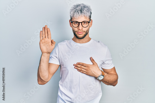 Young hispanic man with modern dyed hair wearing white t shirt and glasses swearing with hand on chest and open palm, making a loyalty promise oath