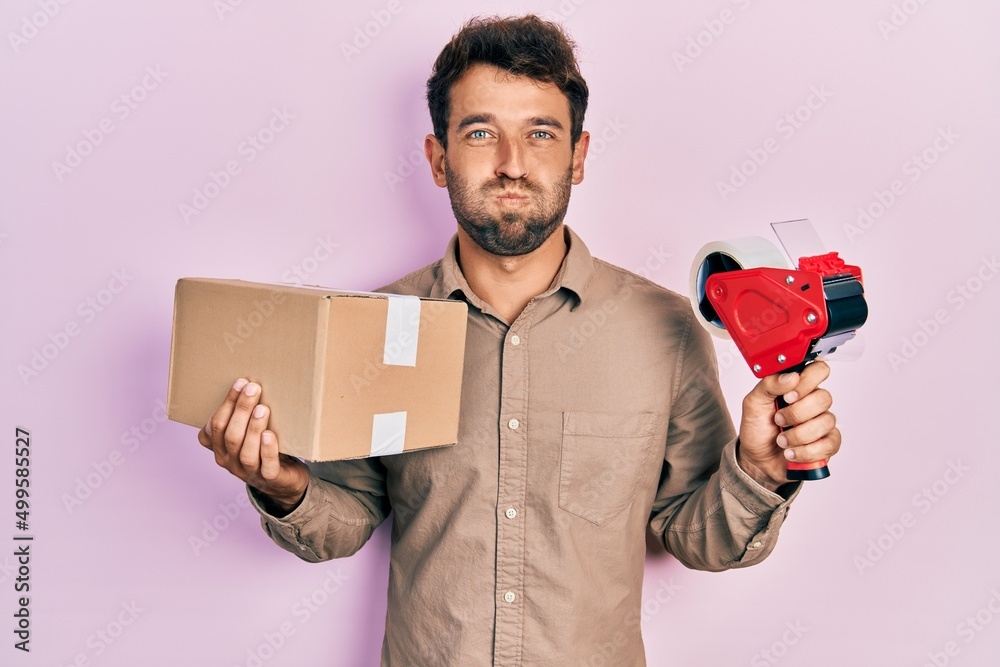 Handsome man with beard holding packing tape holding cardboard puffing cheeks with funny face. mouth inflated with air, catching air. - obrazy, fototapety, plakaty 