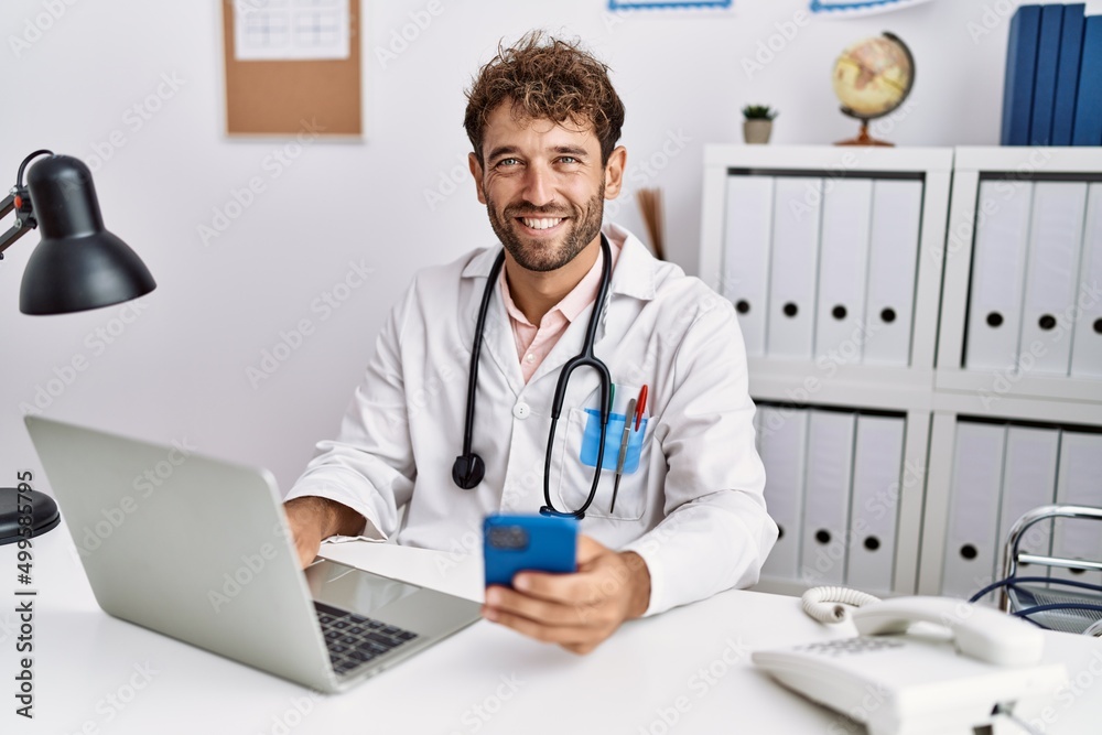Young hispanic man wearing doctor uniform using smartphone at clinic Young hispanic man wearing doctor uniform using smartphone at clinic